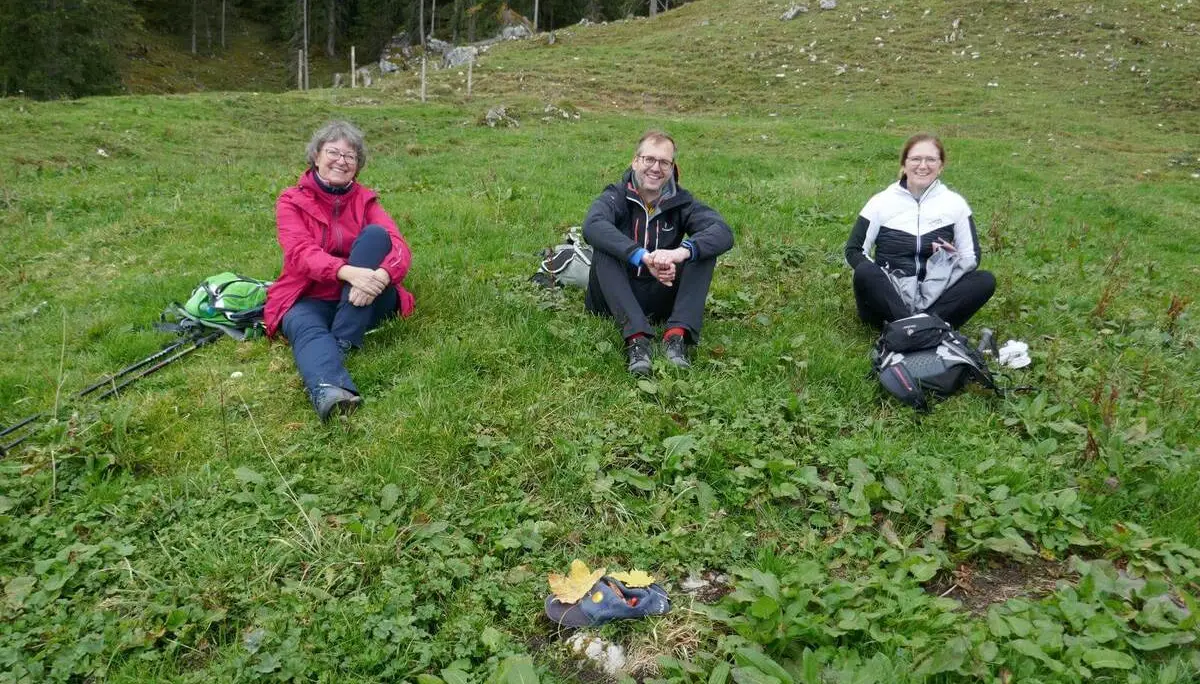 Drei Wandernde sitzen mit Rucksäcken auf grüner Wiese vor Waldrand; im Vordergrund liegt eine Mütze mit Blättern und Naturfundstücken. | © DAV Markt Schwaben/Elke Kressirer