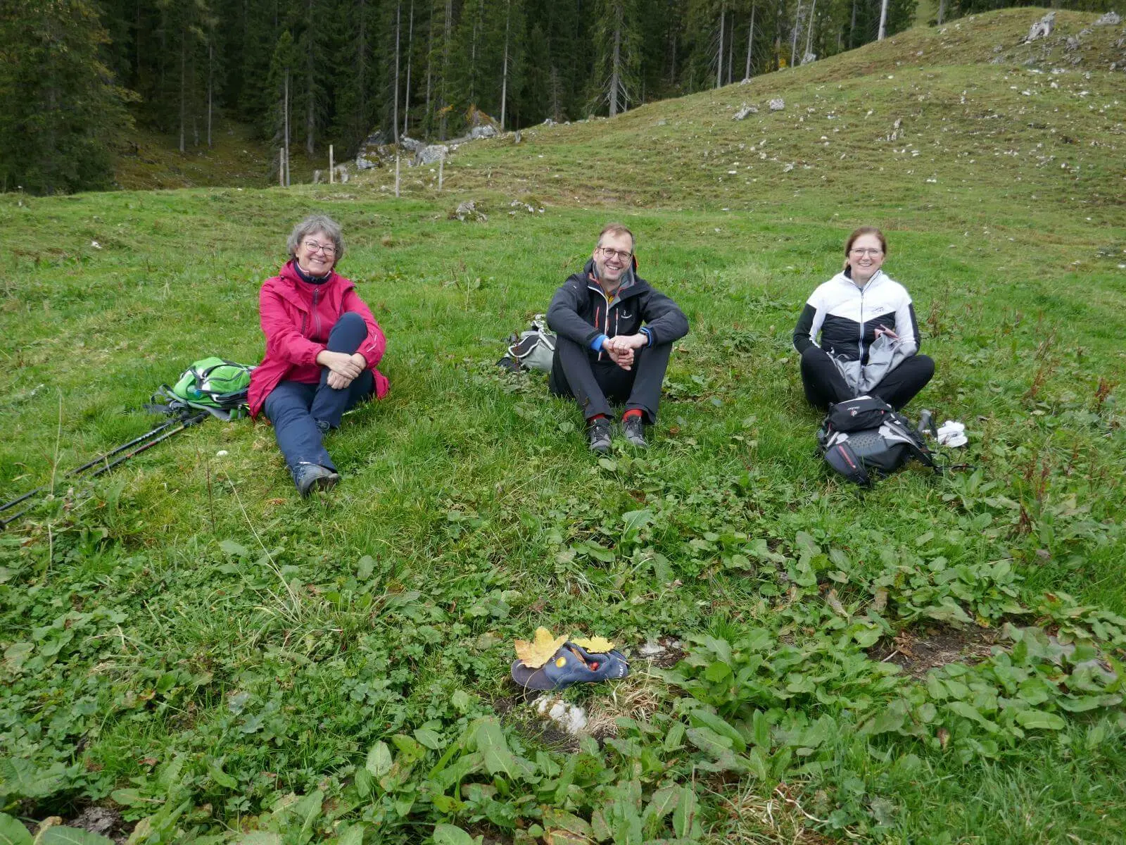 Drei Wandernde sitzen mit Rucksäcken auf grüner Wiese vor Waldrand; im Vordergrund liegt eine Mütze mit Blättern und Naturfundstücken. | © DAV Markt Schwaben/Elke Kressirer