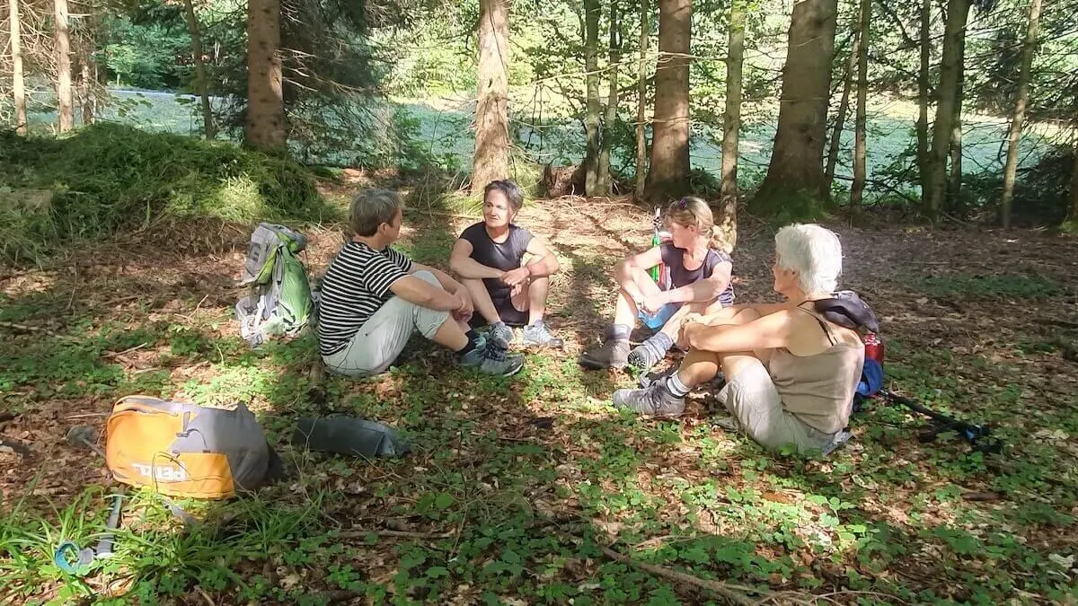 Vier Wandernde sitzen im Wald auf dem Boden, umgeben von hohen Bäumen; Rucksäcke und Stöcke liegen neben ihnen, Sonnenflecken fallen durch das Blätterdach. | © DAV Markt Schwaben/Anna Maria Blau