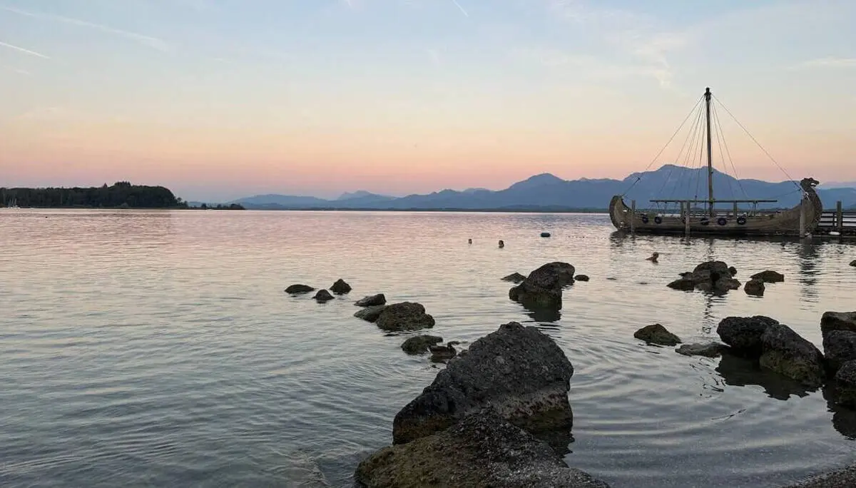 Ruhiger See mit großen Ufersteinen, badenden Menschen und einem hölzernen Boot am Steg; dahinter silhouettierte Berge im warmen Abendlicht. | © DAV Markt Schwaben/Elke Kressirer