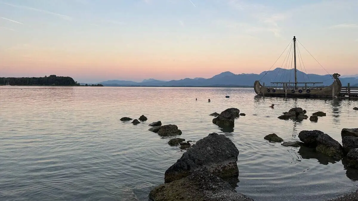 Ruhiger See mit großen Ufersteinen, badenden Menschen und einem hölzernen Boot am Steg; dahinter silhouettierte Berge im warmen Abendlicht. | © DAV Markt Schwaben/Elke Kressirer