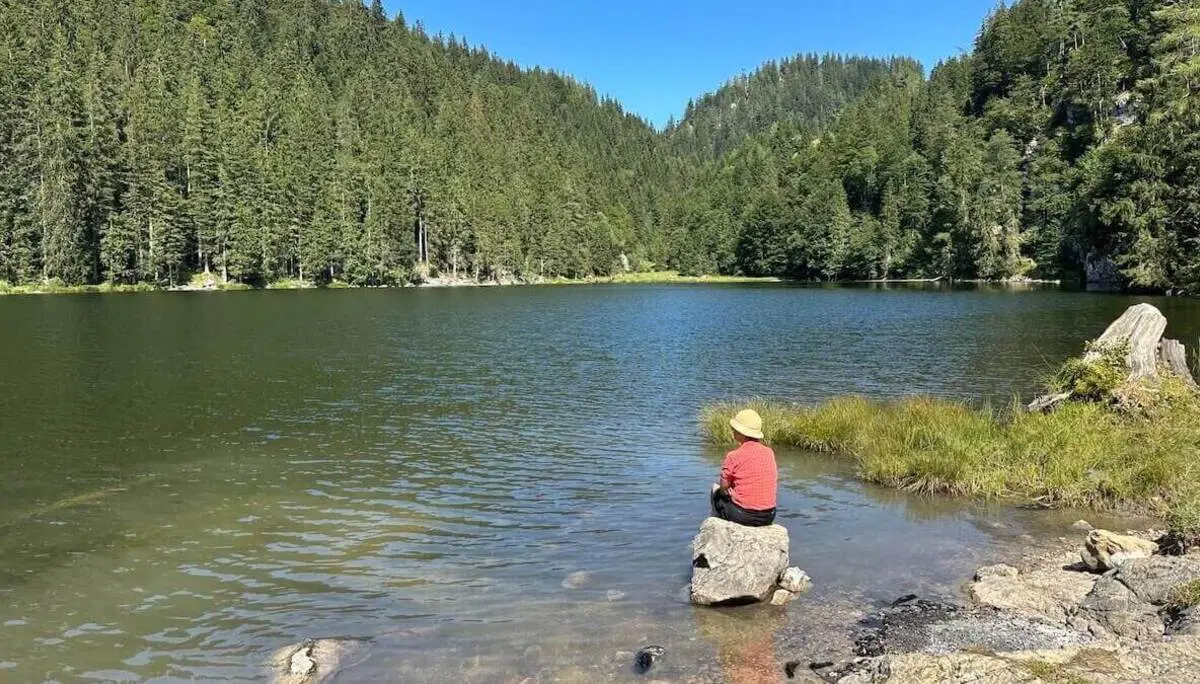 Person mit Strohhut sitzt auf einem Felsen am klaren Bergsee, umgeben von dichtem Nadelwald; ruhige Wasserfläche spiegelt das Grün der Bäume unter blauem Himmel. | © DAV Markt Schwaben/Elke Kressirer