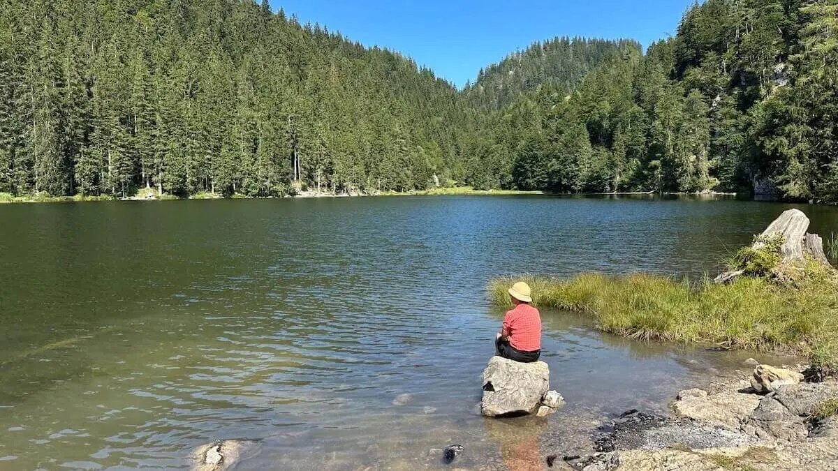Person mit Strohhut sitzt auf einem Felsen am klaren Bergsee, umgeben von dichtem Nadelwald; ruhige Wasserfläche spiegelt das Grün der Bäume unter blauem Himmel. | © DAV Markt Schwaben/Elke Kressirer
