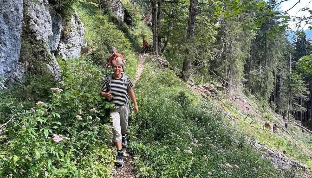 Zwei Wandernde gehen auf einem schmalen Waldweg zwischen Felsen und dichtem Grün; hohe Nadelbäume und üppige Vegetation säumen den sonnigen Bergpfad. | © DAV Markt Schwaben/Elke Kressirer