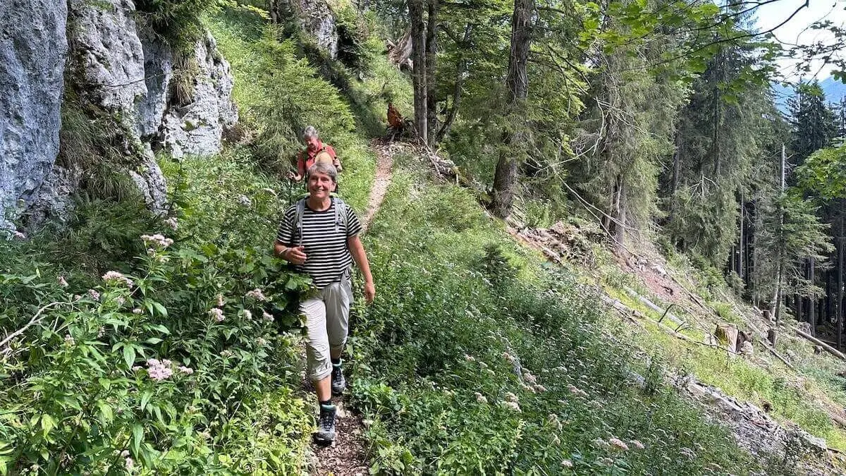 Zwei Wandernde gehen auf einem schmalen Waldweg zwischen Felsen und dichtem Grün; hohe Nadelbäume und üppige Vegetation säumen den sonnigen Bergpfad. | © DAV Markt Schwaben/Elke Kressirer