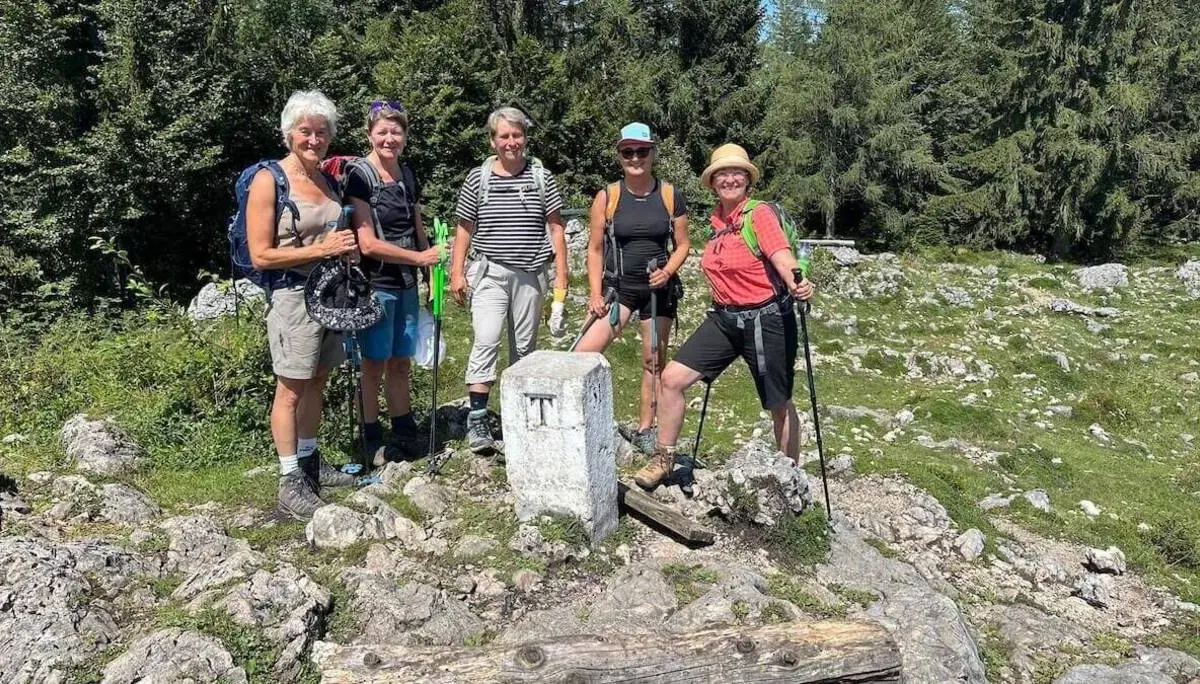 Fünf Wandernde stehen um einen weißen Steinmarker auf felsigem Boden, umgeben von Gras und dichtem Wald bei Sonnenschein. | © DAV Markt Schwaben/Elke Kressirer
