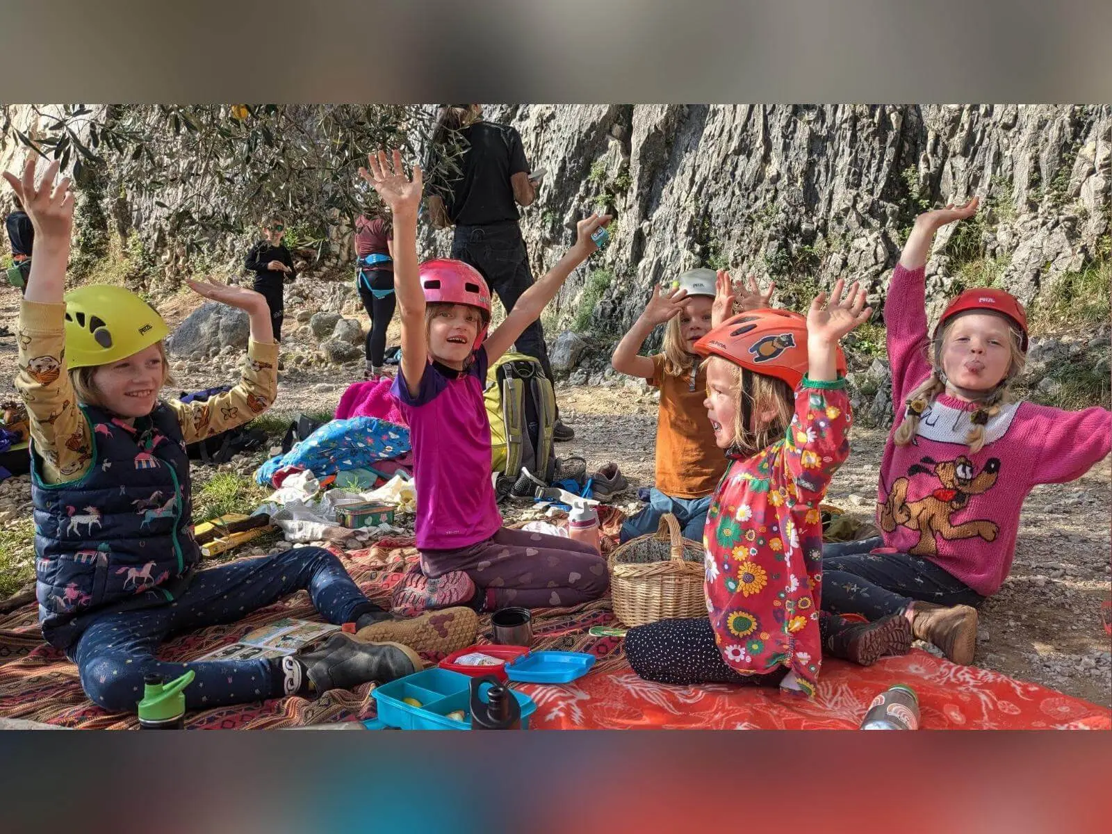 Fünf Kinder mit Kletterhelmen sitzen auf Decken vor einer Felswand in Arco und strecken bei einer Pause die Arme hoch. | © DAV Markt Schwaben / Silke Titze