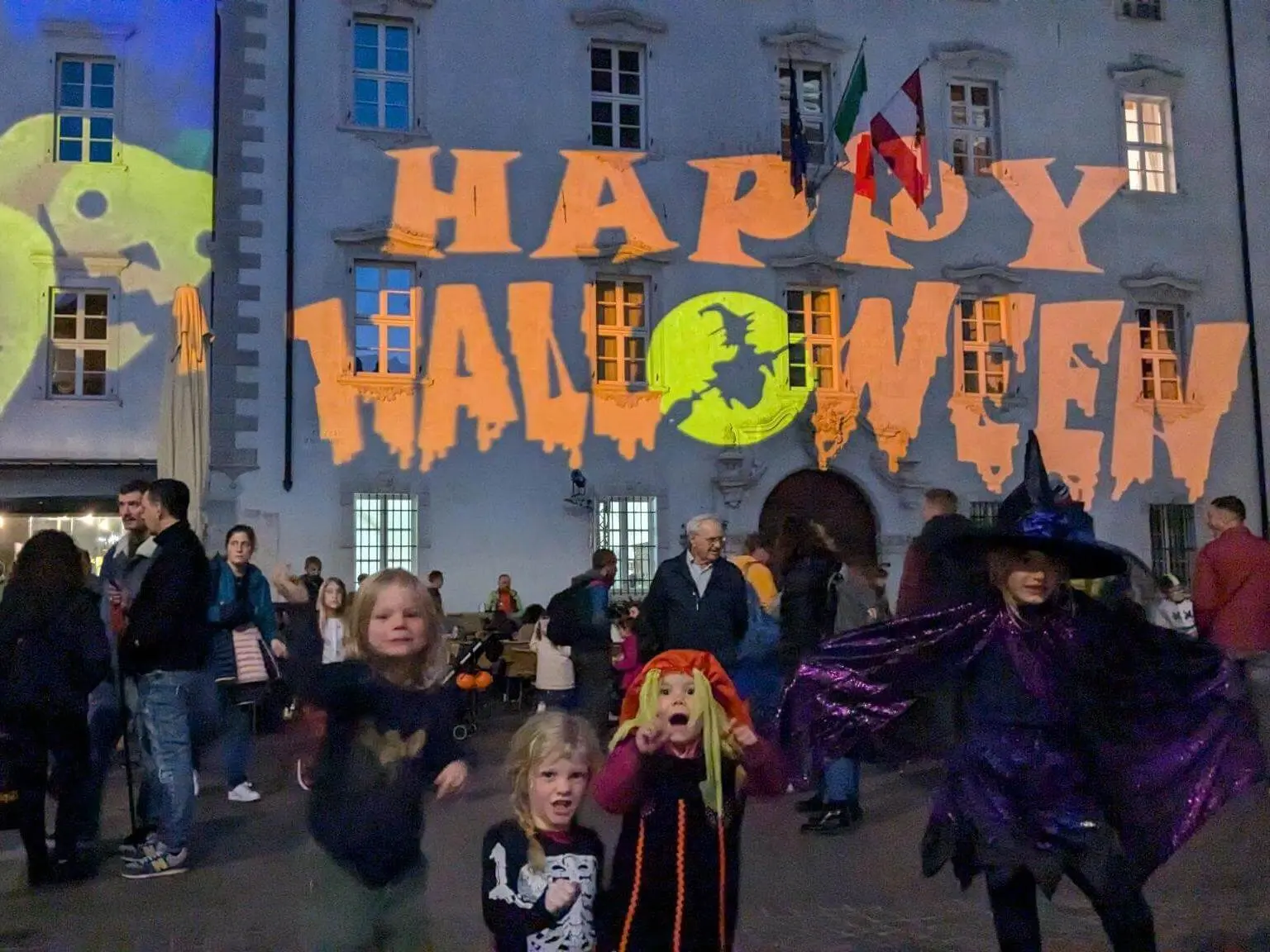 Kinder in Halloweenkostümen stehen auf einem Platz vor einem Gebäude mit großer „Happy Halloween“-Projektion und vielen Besuchern. | © DAV Markt Schwaben / Silke Titze