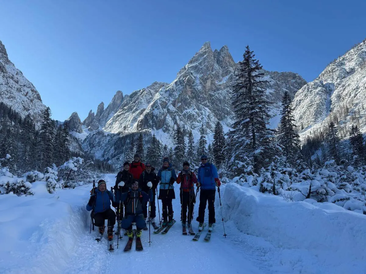 Acht Skitourengeher:innen stehen auf verschneitem Waldpfad im Fischleinbachtal vor sonnenbeschienenen Dolomitengipfeln. | © DAV Markt Schwaben · Foto: Benedikt Scheuerecke