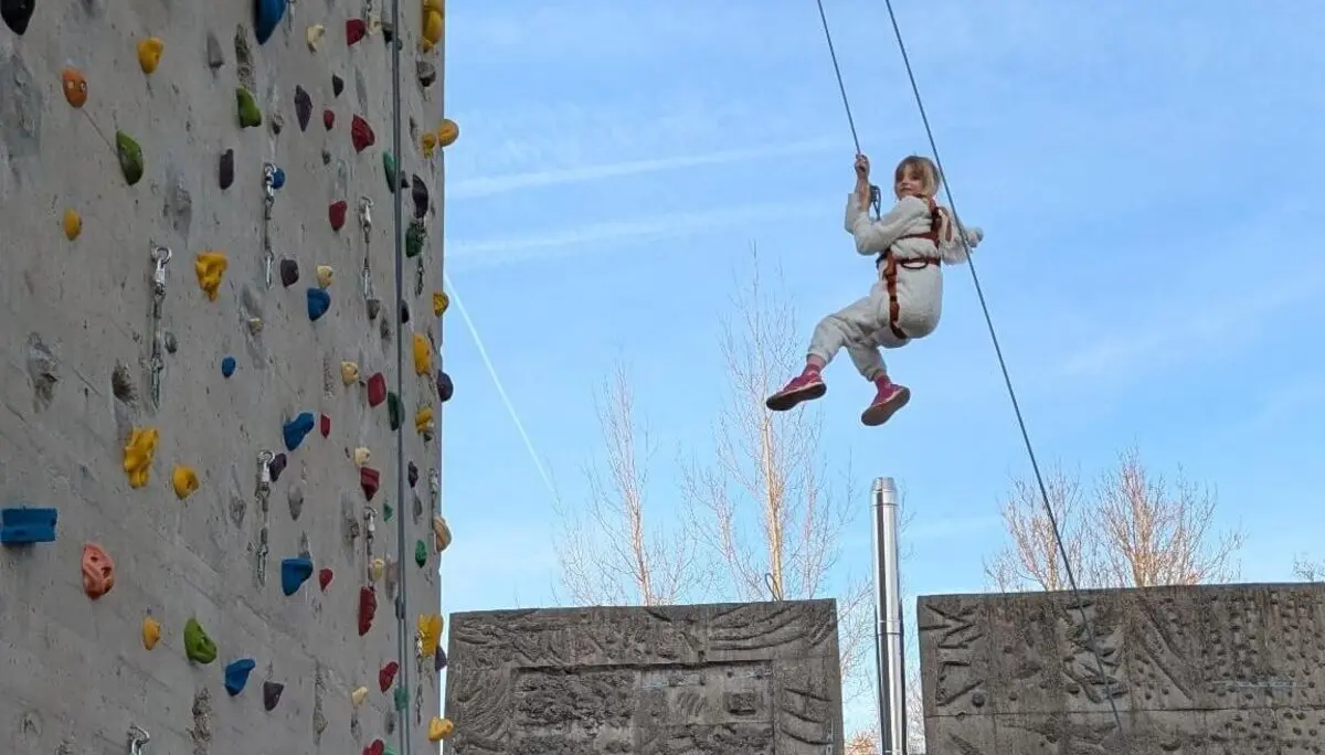 Ein junges Mädchen in einem weißen Overall und rosa Schuhen hängt in der Luft, während es sich von einer hohen Kletterwand im Freien mit bunten Griffen abseilt. | © DAV Markt Schwaben