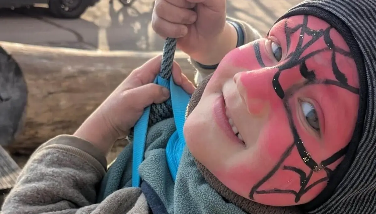 Ein lächelndes Kind mit einer rot-schwarzen Spider-Man-Gesichtsbemalung hält sich an einem Seil fest, während es im Freien schaukelt, bekleidet mit einer warmen Jacke und einer Mütze. | © DAV Markt Schwaben