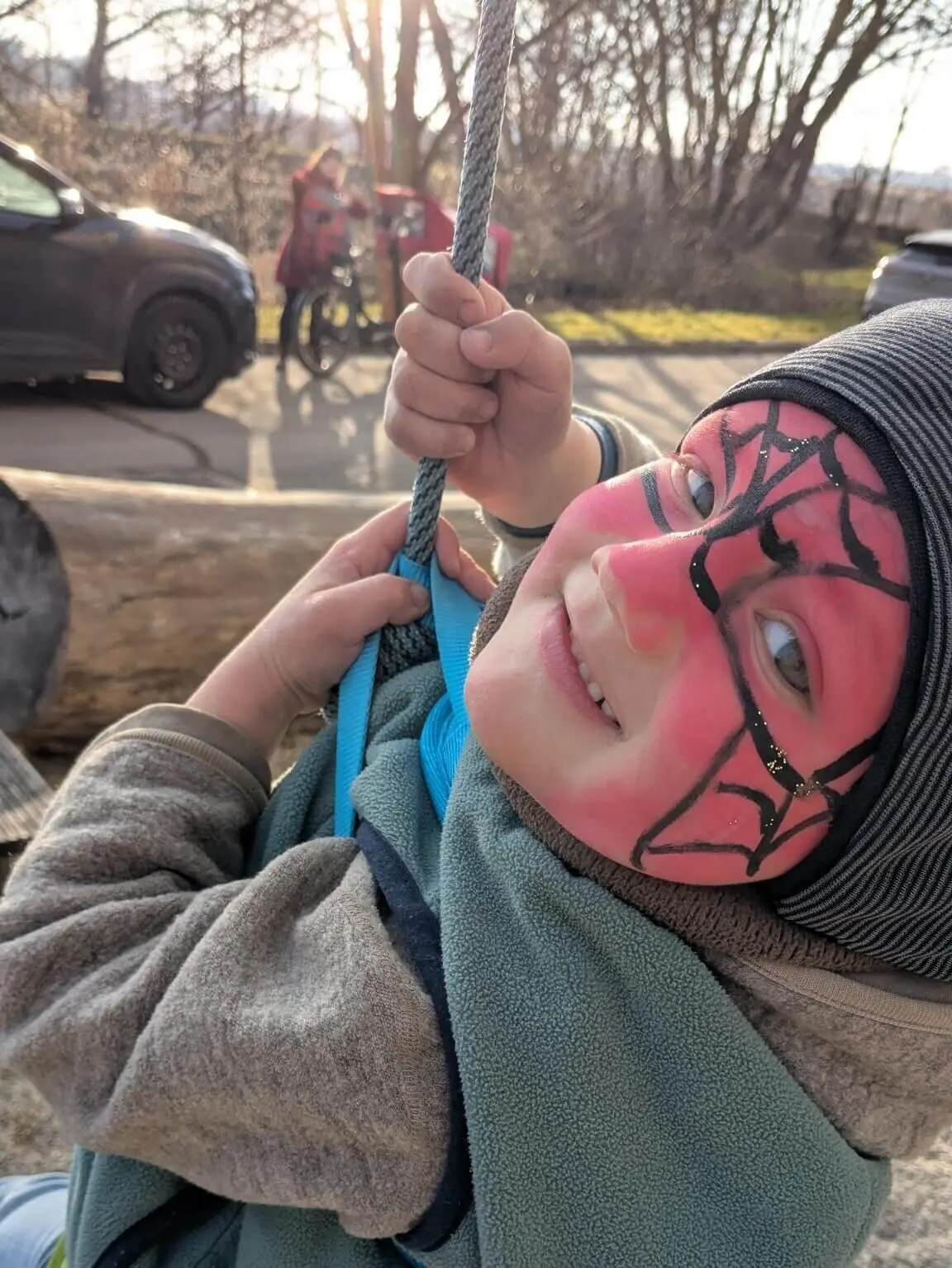 Ein lächelndes Kind mit einer rot-schwarzen Spider-Man-Gesichtsbemalung hält sich an einem Seil fest, während es im Freien schaukelt, bekleidet mit einer warmen Jacke und einer Mütze. | © DAV Markt Schwaben