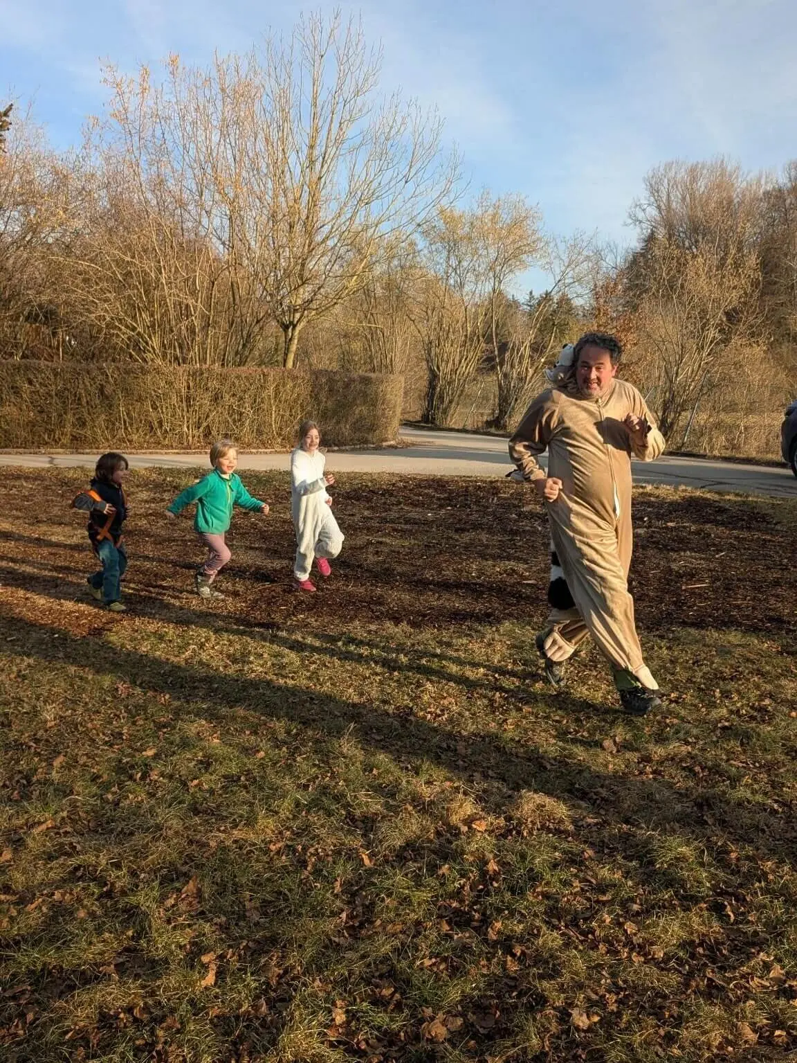 Eine Gruppe von vier Kindern und einem Erwachsenen rennt draußen auf einem mit Laub bedeckten Boden. Der Erwachsene trägt ein Kostüm, das wie ein Tier aussieht. Die Kinder tragen verschiedene farbenfrohe Kleidung. Im Hintergrund sind kahle Bäume und ein Weg zu sehen. Die Szene findet an einem sonnigen Tag statt. | © DAV Markt Schwaben
