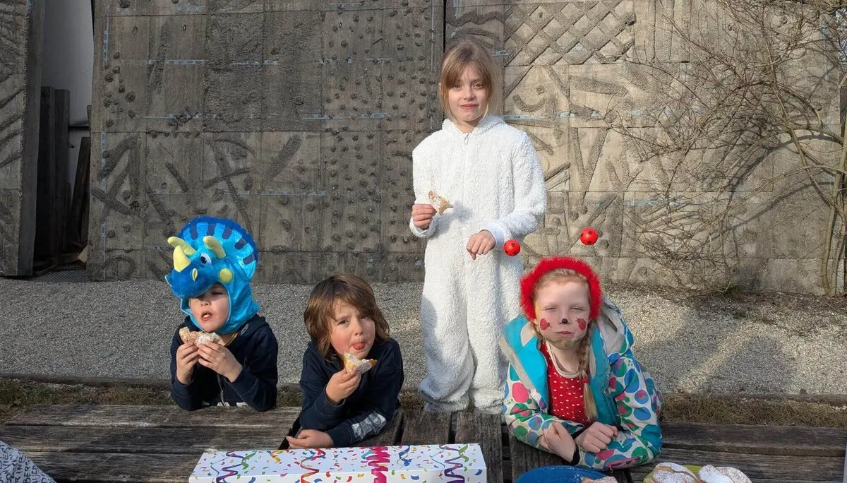 Vier Kinder in bunten und verspielten Outfits sitzen draußen an einem Holztisch und essen Krampfen. | © DAV Markt Schwaben