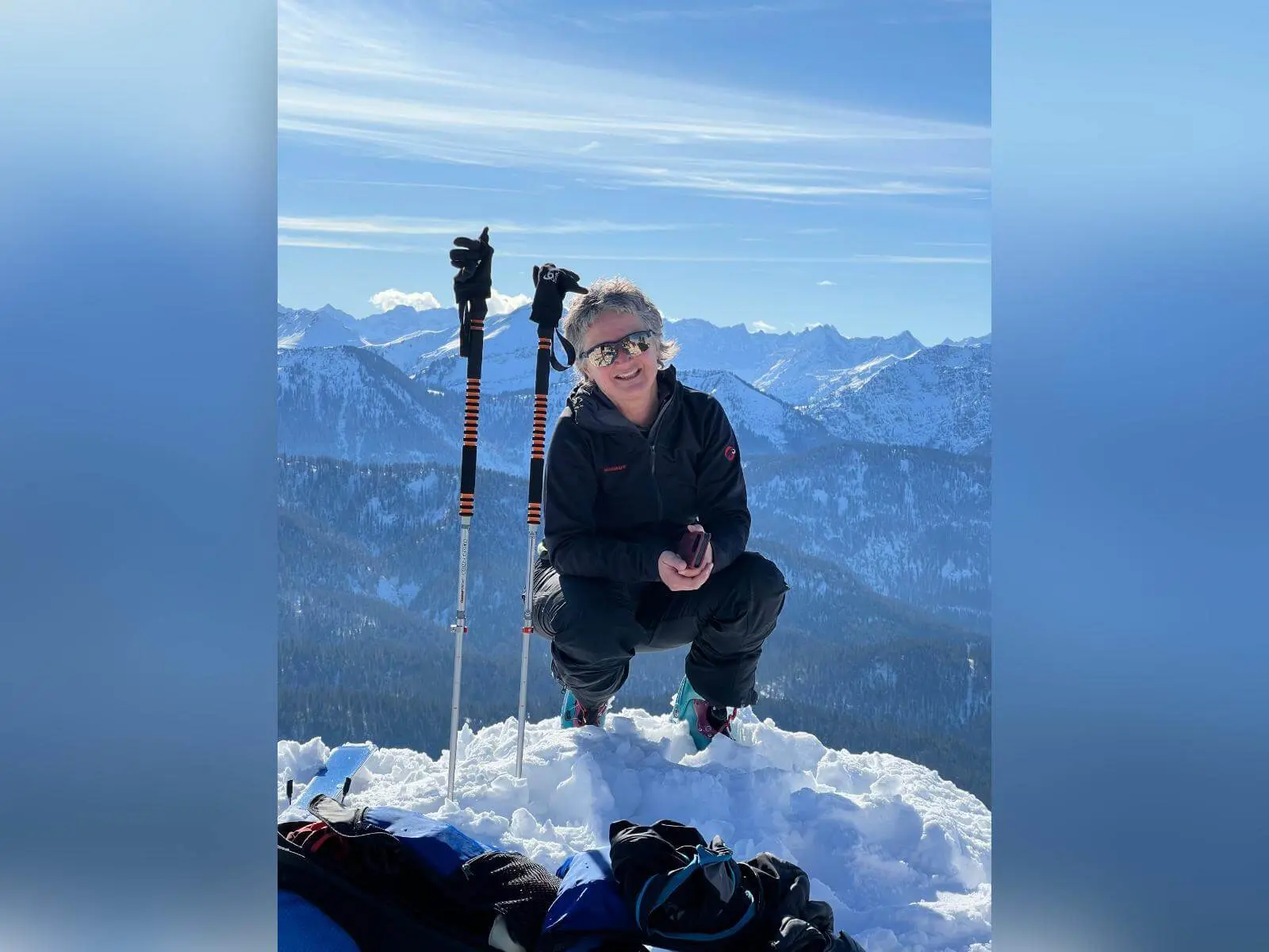 Person kniet im Schnee auf einem Gipfel neben Skistöcken, im Hintergrund erstreckt sich ein weites, sonniges Bergpanorama.Provide your feedback on BizChat | © DAV Markt Schwaben/Elke Kressirer