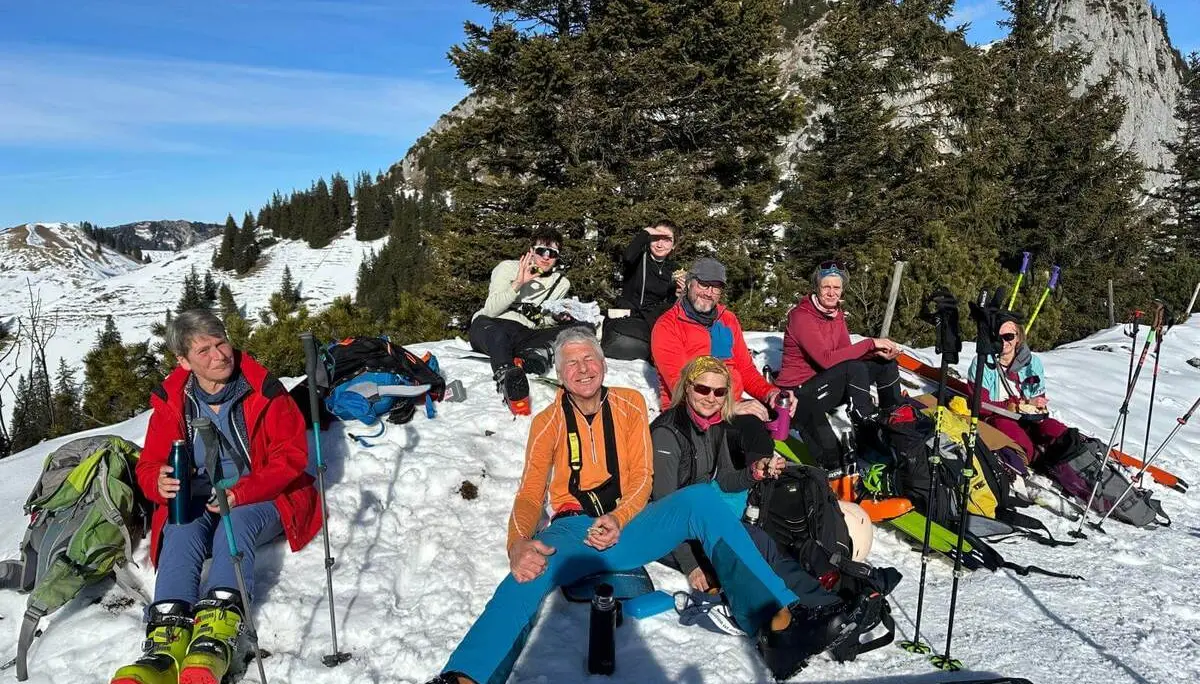 Mehrere Personen sitzen im Schnee bei einer Pause, umgeben von Skiern, Stöcken und Rucksäcken; dahinter liegen verschneite Berge und Bäume. | © DAV Markt Schwaben/Elke Kressirer