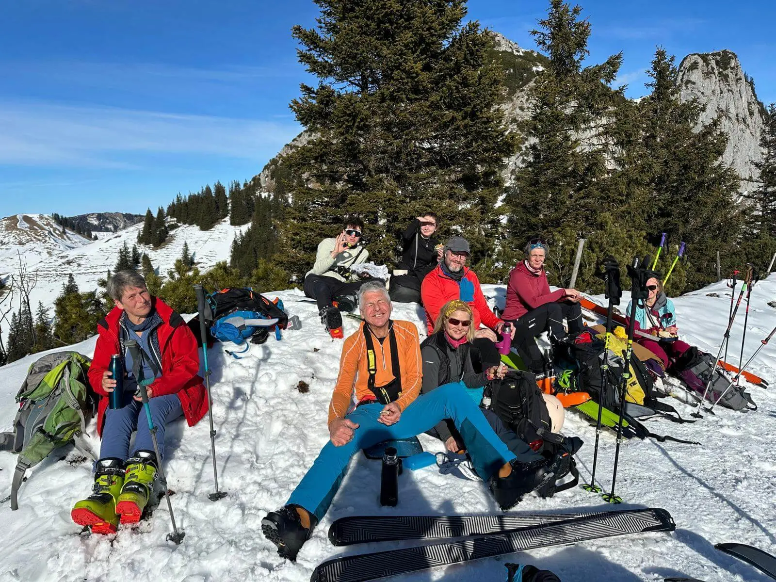 Mehrere Personen sitzen im Schnee bei einer Pause, umgeben von Skiern, Stöcken und Rucksäcken; dahinter liegen verschneite Berge und Bäume. | © DAV Markt Schwaben/Elke Kressirer