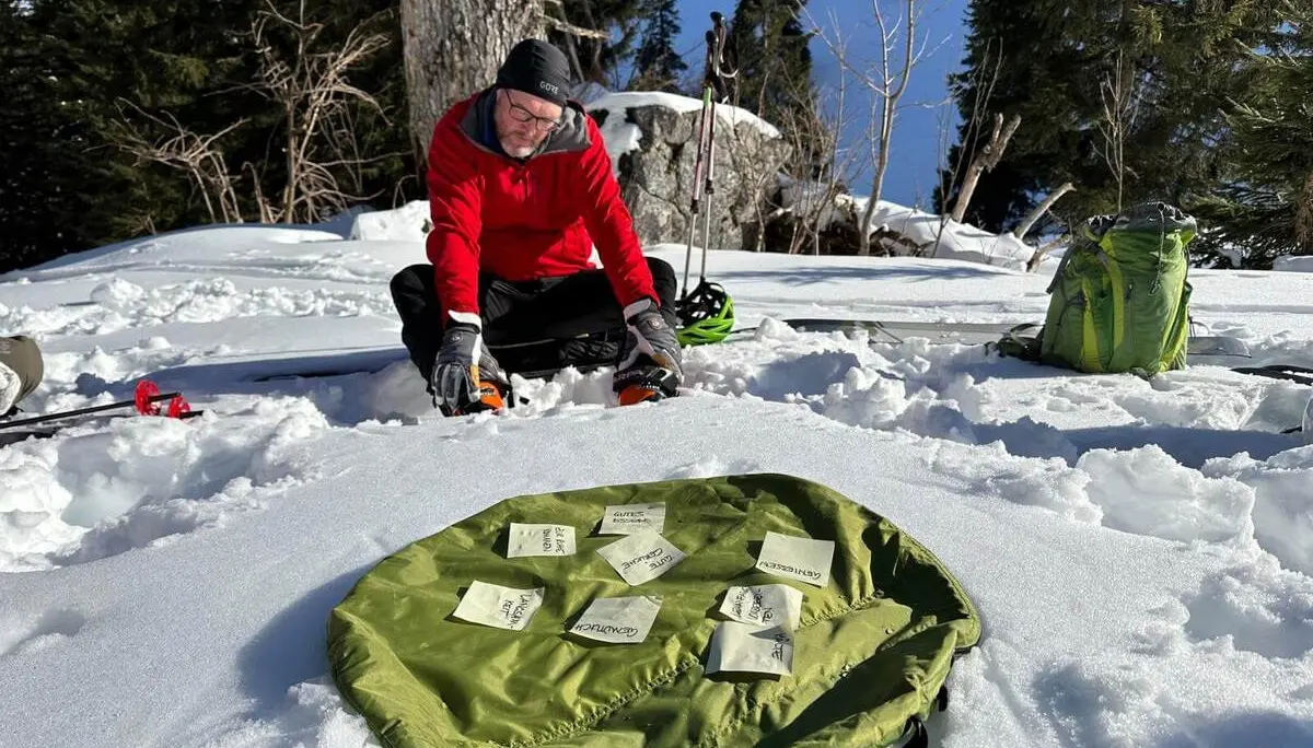 Person kniet im Schnee vor ausgebreitetem Rucksack mit beschrifteten Zetteln; dahinter stehen Skistöcke und Bäume in winterlicher Berglandschaft. | © DAV Markt Schwaben/Elke Kressirer