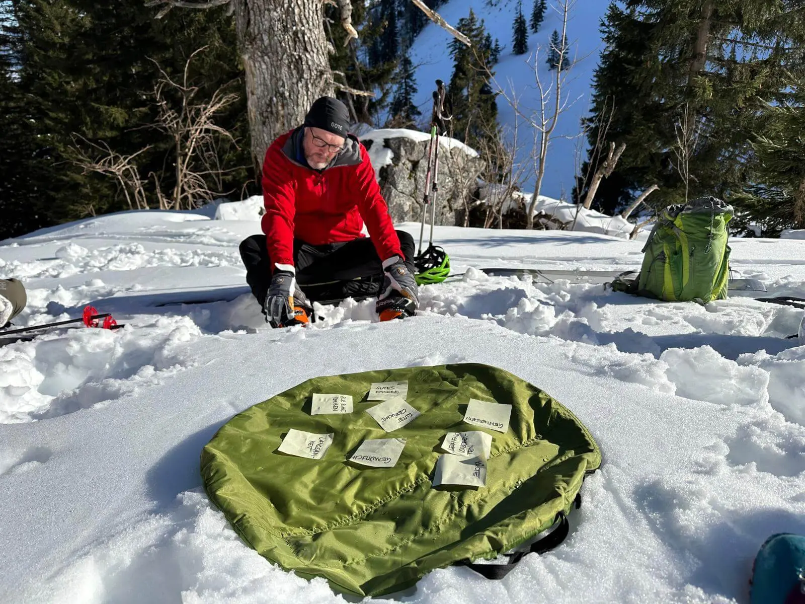Person kniet im Schnee vor ausgebreitetem Rucksack mit beschrifteten Zetteln; dahinter stehen Skistöcke und Bäume in winterlicher Berglandschaft. | © DAV Markt Schwaben/Elke Kressirer