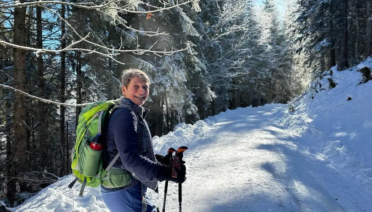 Person mit Rucksack und Stöcken steht auf verschneitem Weg im Wald; schneebedeckte Bäume und Sonnenlicht prägen die winterliche Szene. | © DAV Markt Schwaben/Elke Kressirer
