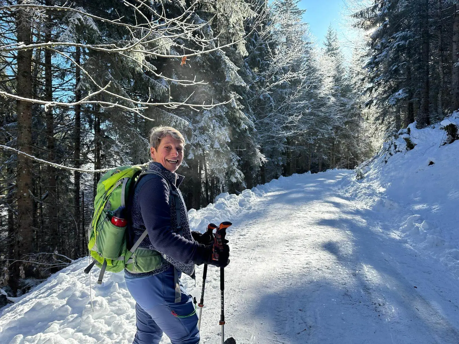 Person mit Rucksack und Stöcken steht auf verschneitem Weg im Wald; schneebedeckte Bäume und Sonnenlicht prägen die winterliche Szene. | © DAV Markt Schwaben/Elke Kressirer