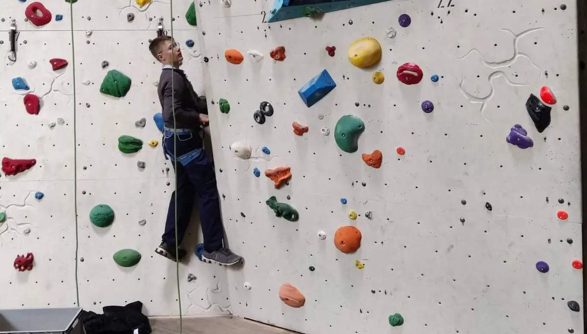 Eine Person klettert gesichert an einer bunten Indoor-Kletterwand, Kletterausrüstung liegt am Boden. | © DAV Markt Schwaben