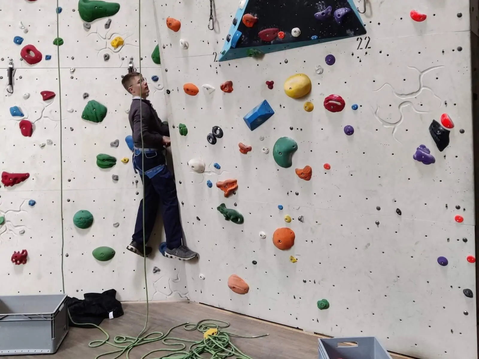 Eine Person klettert gesichert an einer bunten Indoor-Kletterwand, Kletterausrüstung liegt am Boden. | © DAV Markt Schwaben