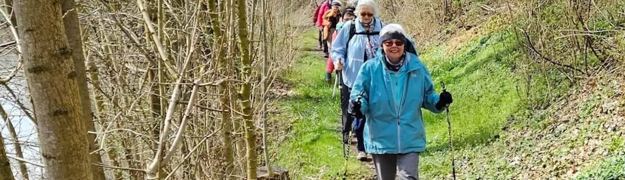 Seniorengruppe beim Wandern auf einem schmalen Waldpfad. Die Wanderer tragen wetterfeste Kleidung und nutzen Wanderstöcke. Umgeben von kahlen Bäumen und grünem Unterholz deutet die Szenerie auf den frühen Frühling hin. Die Gruppe bewegt sich in einer Reihe entlang des naturnahen Weges. | © DAV Markt Schwaben