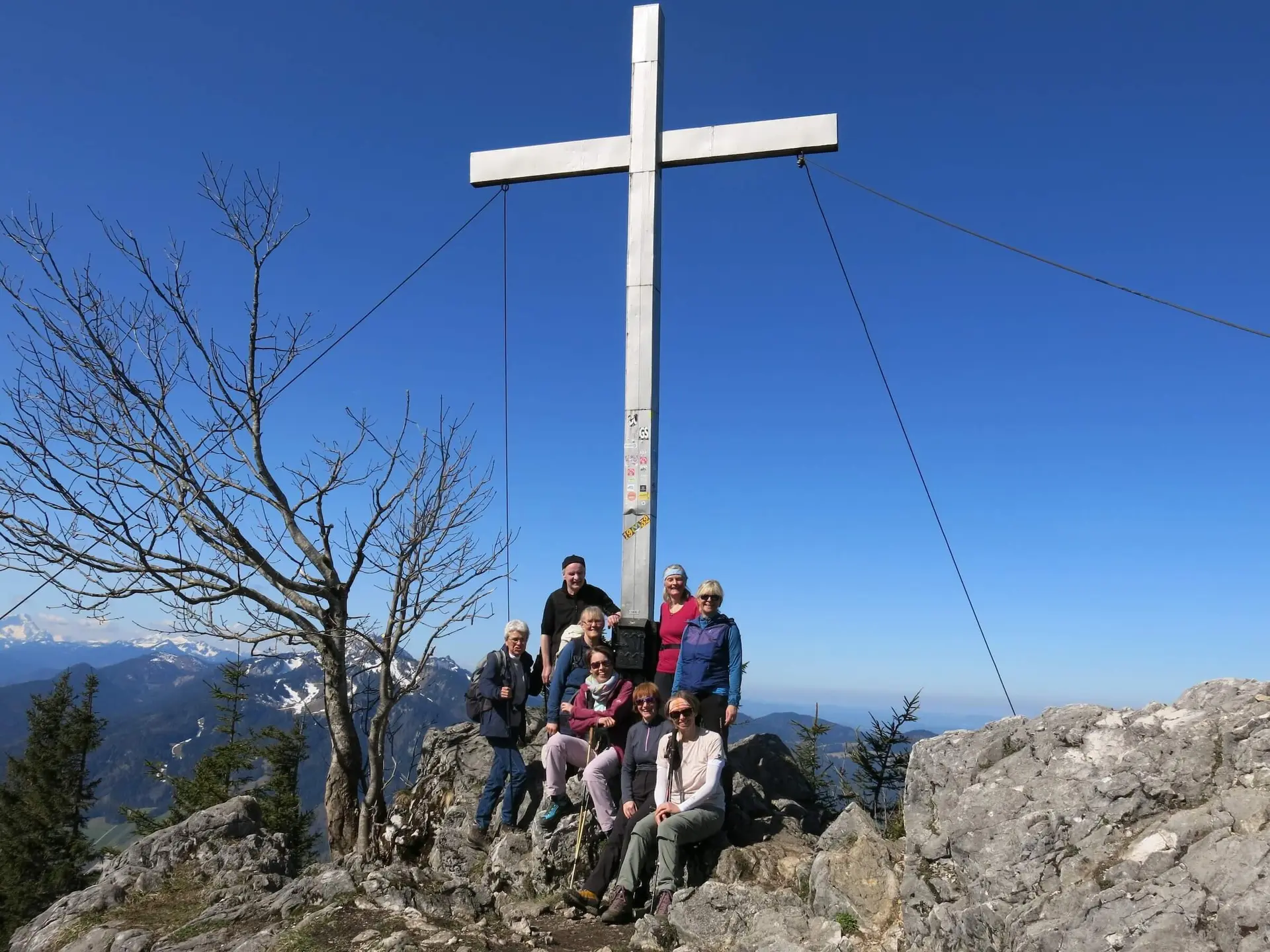 Gruppe von Wanderern auf felsigem Gipfel neben einem großen Metallkreuz. Klarer Himmel und Alpenpanorama im Hintergrund sorgen für eine beeindruckende Aussicht. | © DAV Markt Schwaben | Foto Erwin Matzinger