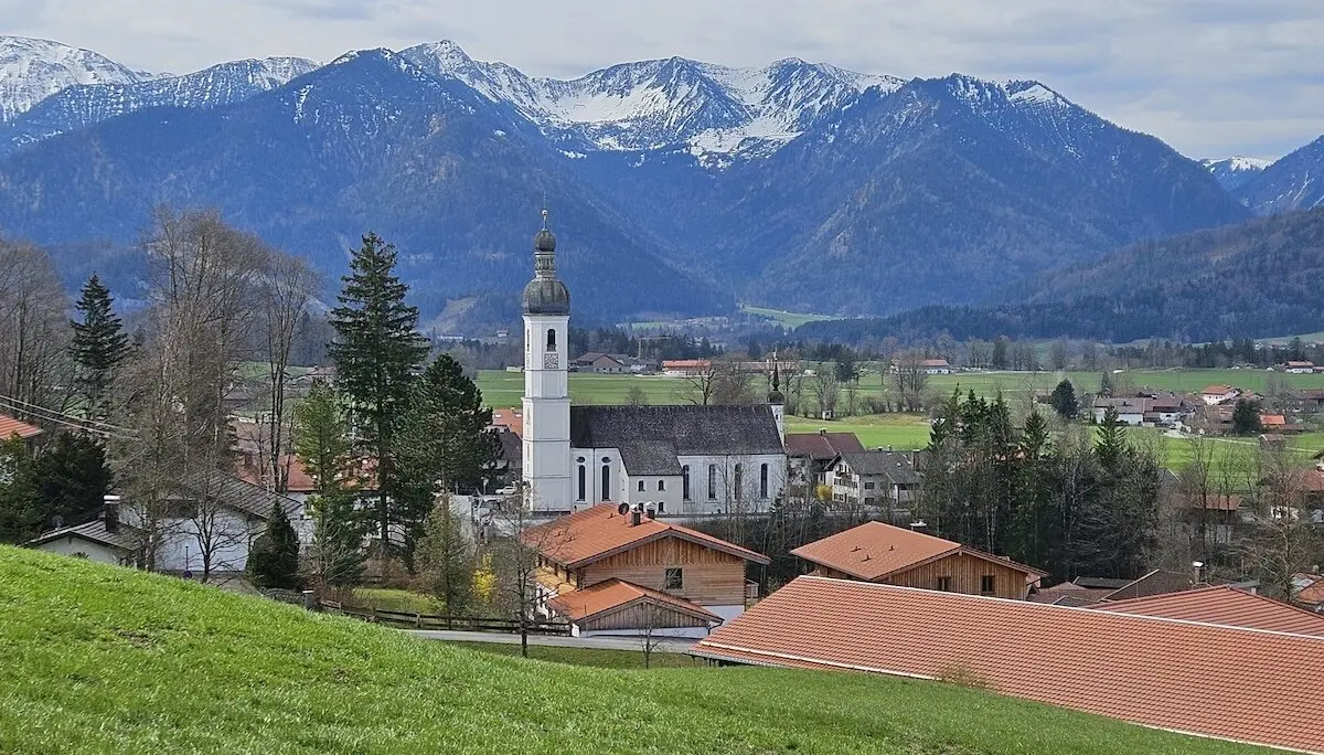 Malerisches Dorf im Tal mit schneebedeckten Bergen im Hintergrund, weißer Kirche mit grüner Zwiebelturmspitze und roten Hausdächern, umgeben von grünen Feldern und Bäumen. | © DAV Markt Schwaben | Foto Hubert Inhofer