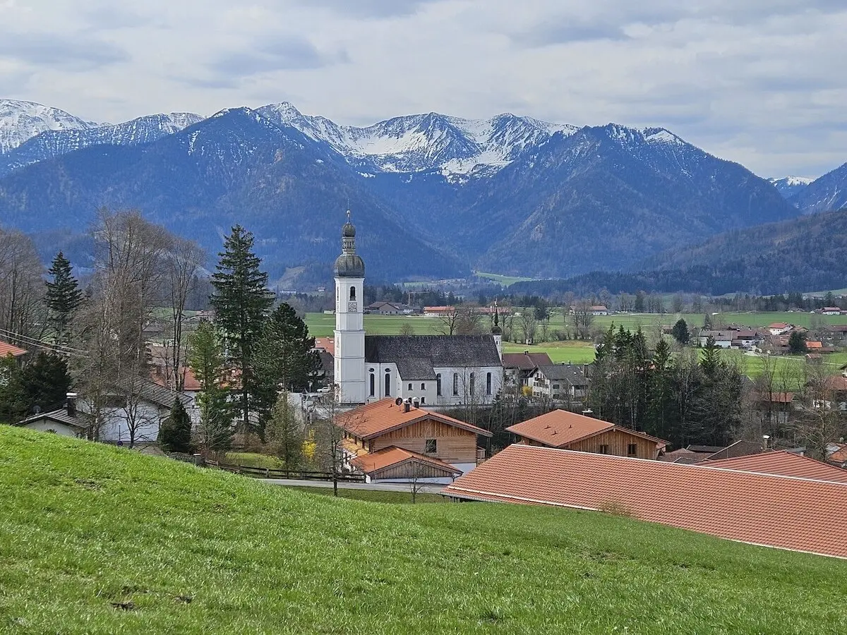 Malerisches Dorf im Tal mit schneebedeckten Bergen im Hintergrund, weißer Kirche mit grüner Zwiebelturmspitze und roten Hausdächern, umgeben von grünen Feldern und Bäumen. | © DAV Markt Schwaben | Foto Hubert Inhofer