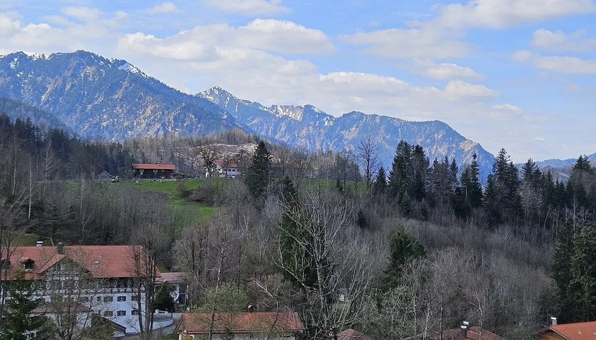 Gemütliches Bergdorf mit roten Dächern, umgeben von Bäumen und grünen Hügeln, während schneebedeckte Gipfel den Horizont prägen. Ein idyllisches und ruhiges Landschaftsbild. | © DAV Markt Schwaben | Foto Hubert Inhofer