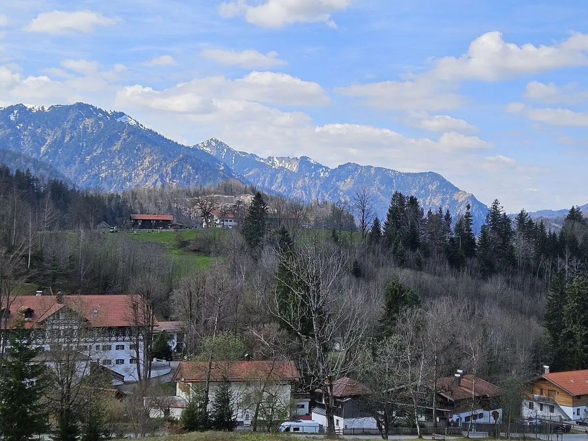 Gemütliches Bergdorf mit roten Dächern, umgeben von Bäumen und grünen Hügeln, während schneebedeckte Gipfel den Horizont prägen. Ein idyllisches und ruhiges Landschaftsbild. | © DAV Markt Schwaben | Foto Hubert Inhofer