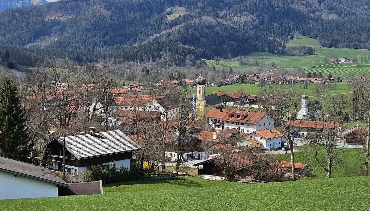 Malerisches Dorf in einem Tal mit roten Ziegeldächern, einer Kirche mit gelbem Glockenturm und grünen Wiesen im Vordergrund, umgeben von bewaldeten Hügeln und majestätischen Bergen. Ein idyllischer Anblick ländlicher Schönheit. | © DAV Markt Schwaben | Foto Hubert Inhofer
