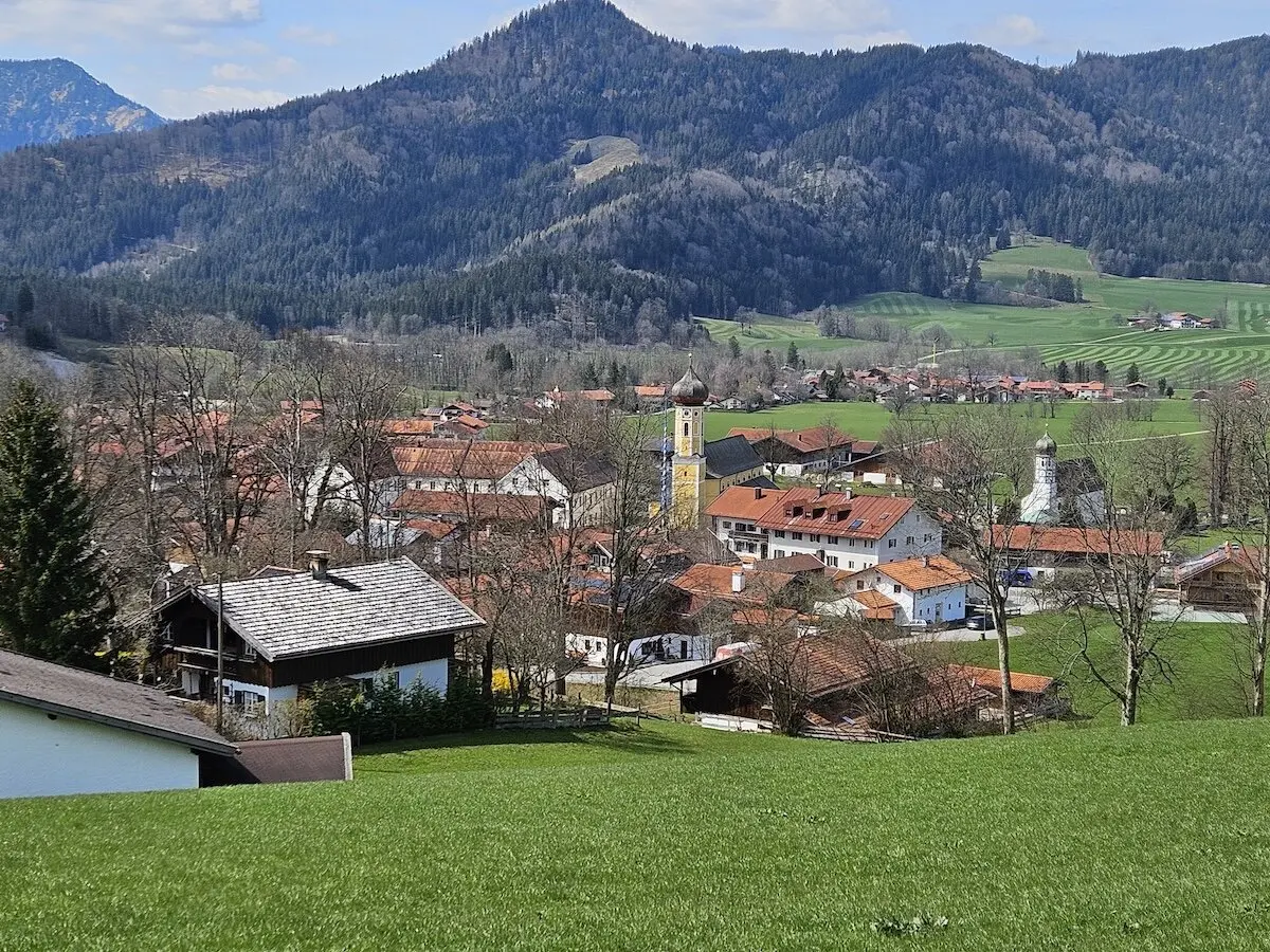 Malerisches Dorf in einem Tal mit roten Ziegeldächern, einer Kirche mit gelbem Glockenturm und grünen Wiesen im Vordergrund, umgeben von bewaldeten Hügeln und majestätischen Bergen. Ein idyllischer Anblick ländlicher Schönheit. | © DAV Markt Schwaben | Foto Hubert Inhofer