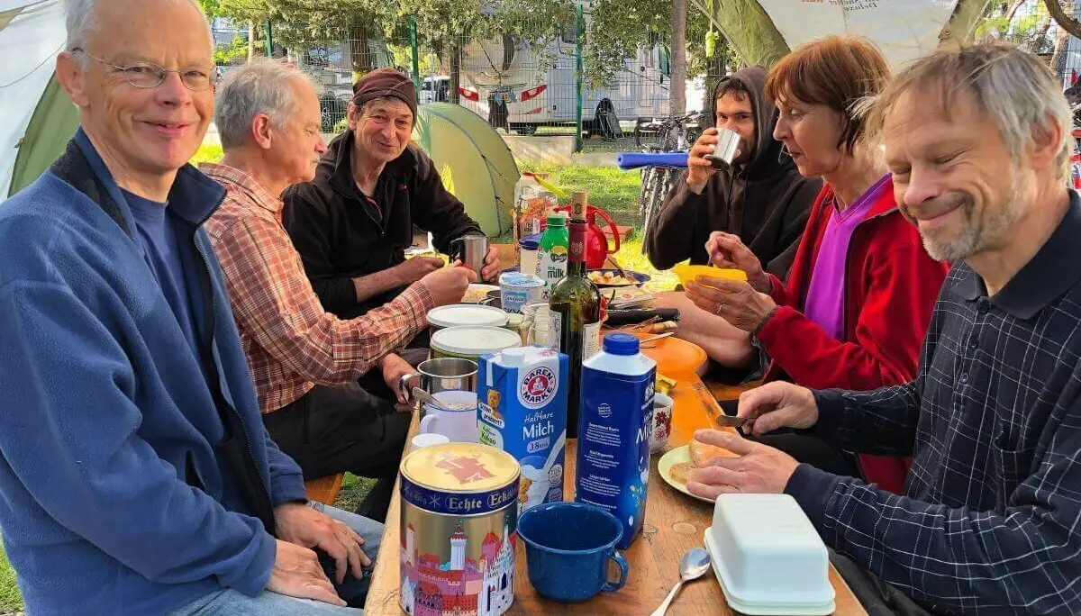 Acht Erwachsene sitzen am Picknicktisch unter einem Pavillon, umgeben von Zelten und Bäumen, beim gemeinsamen Frühstück. | © DAV Markt Schwaben | Foto Hildegard Petschik
