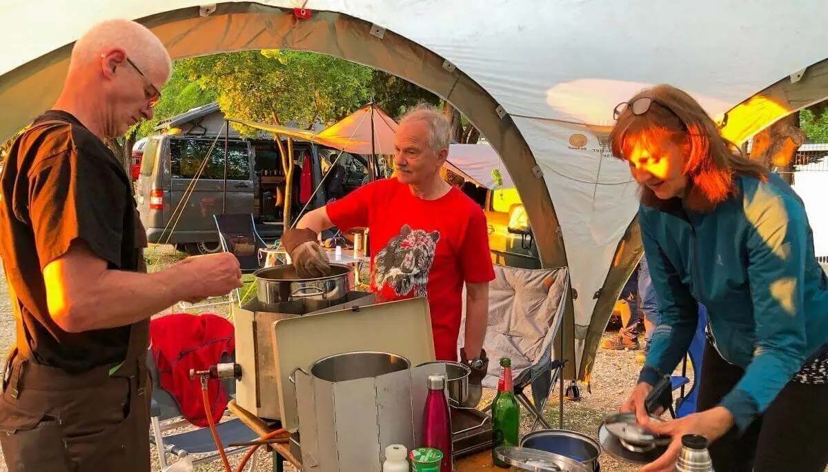 Drei Personen bereiten gemeinsam Essen unter einem Pavillon zu; Campingausrüstung und Zelte im Hintergrund. | © DAV Markt Schwaben | Foto Hildegard Petschik