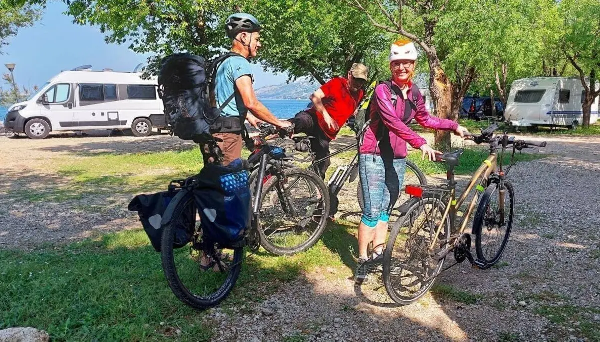 Drei Personen mit Fahrrädern stehen auf einem Campingplatz nahe Campervans; Bäume und Wasser im Hintergrund. | © DAV Markt Schwaben | Foto Lutz Gründel