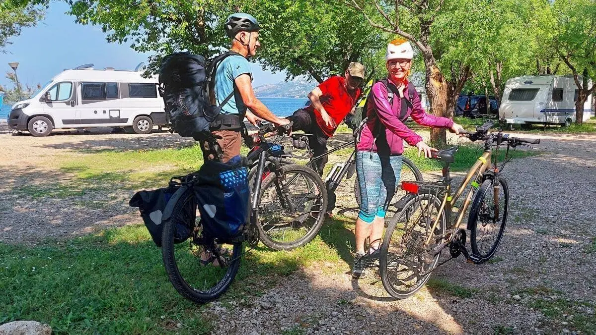 Drei Personen mit Fahrrädern stehen auf einem Campingplatz nahe Campervans; Bäume und Wasser im Hintergrund. | © DAV Markt Schwaben | Foto Lutz Gründel