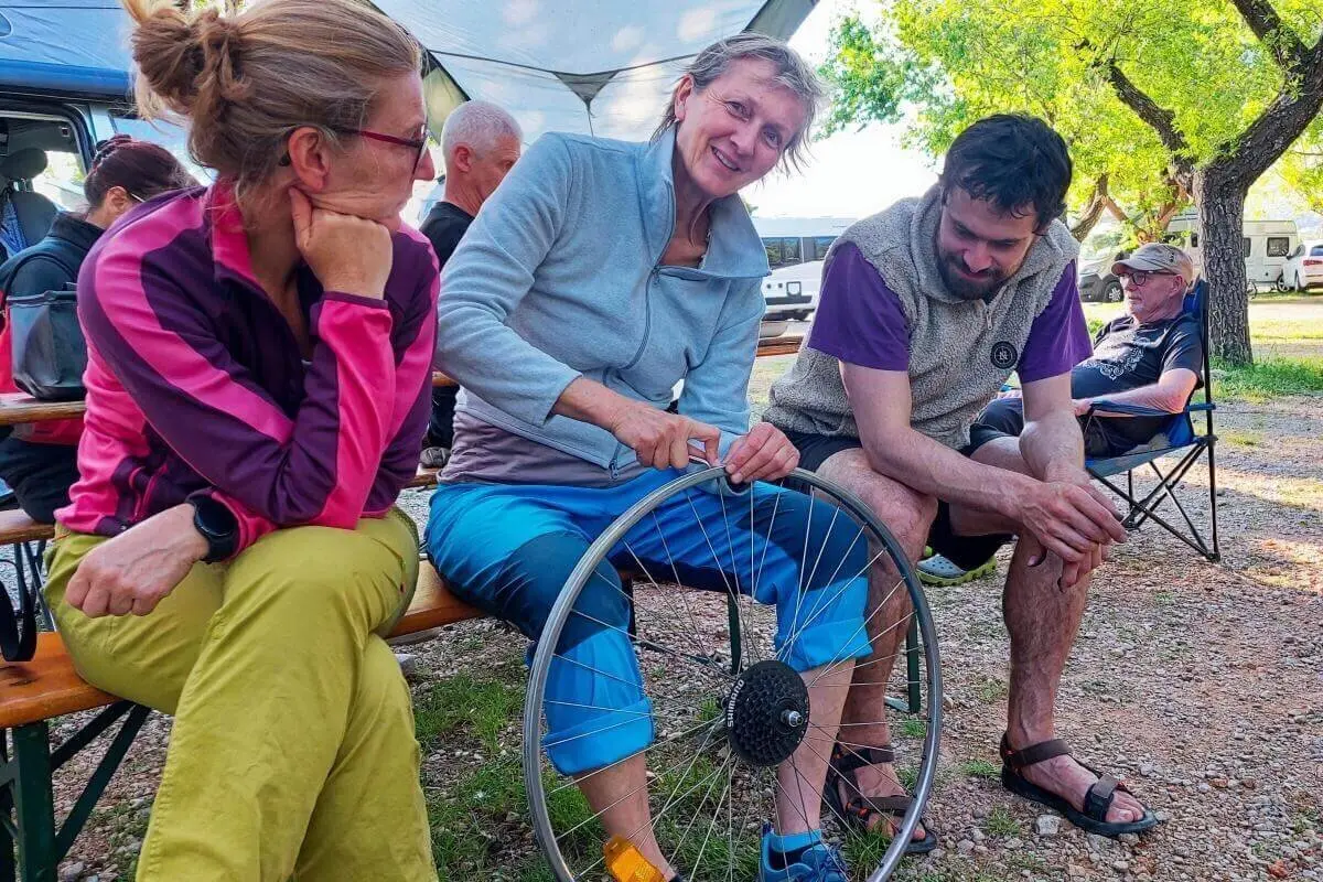 Eine Person zeigt ein Fahrradteil in einer Gruppe; Campervans und Bäume rahmen die Szene. | © DAV Markt Schwaben | Foto Lutz Gründel
