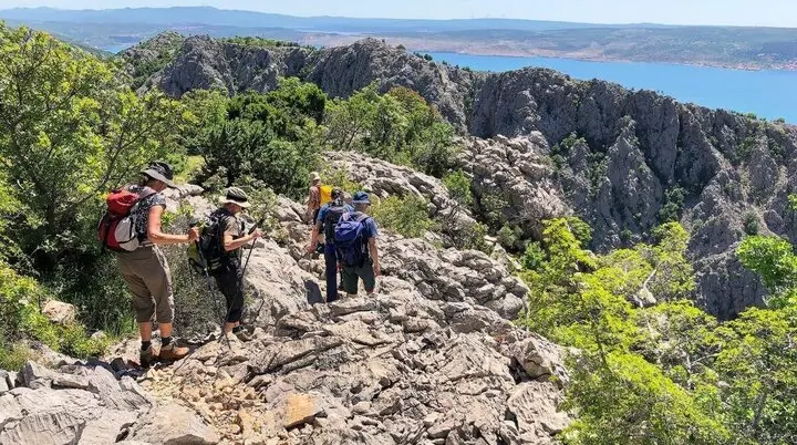Fünf Wandernde auf felsigem Pfad mit Blick auf das Meer und entfernte Berge unter blauem Himmel. | © DAV Markt Schwaben | Foto Hildegard Petschik
