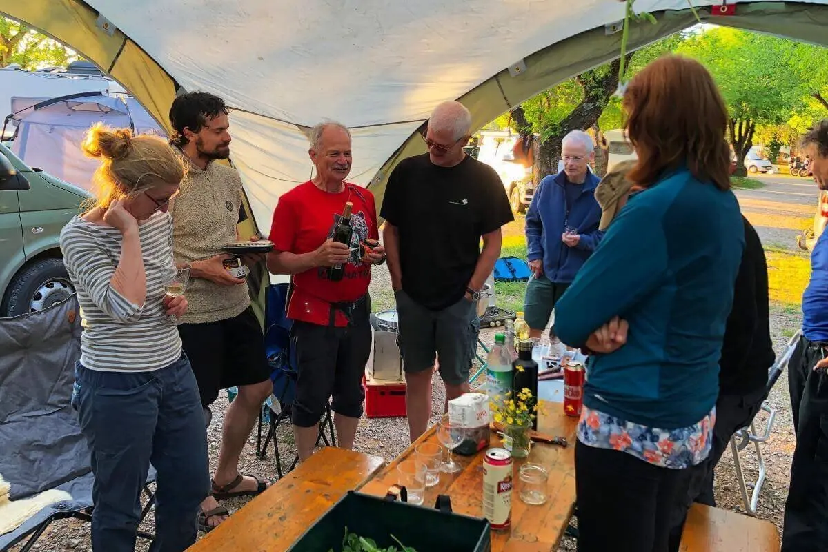 Acht Personen stehen oder sitzen am Tisch mit Getränken und Pflanze; Zelte und Van im Hintergrund. | © DAV Markt Schwaben | Foto Hildegard Petschik