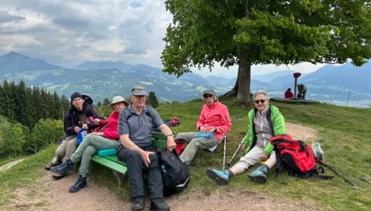 Seniorengruppe macht Pause auf Holzbank mit Bergblick in Oberaudorf. Wolkiger Himmel, entspannte Stimmung. | © DAV Markt Schwaben / Hubert Inhofer