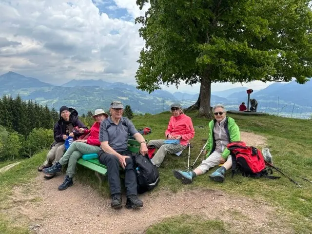 Seniorengruppe macht Pause auf Holzbank mit Bergblick in Oberaudorf. Wolkiger Himmel, entspannte Stimmung. | © DAV Markt Schwaben / Hubert Inhofer
