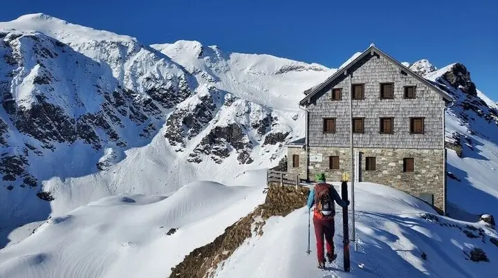 Ein Bergsteiger sitzt mit Ausrüstung neben dem Niedersachsenhaus auf schneebedecktem Hang – im Sonnenlicht eröffnet sich der Blick auf die Schareck-Nordwand. | © DAV Markt Schwaben | Foto: Walter Kressirer