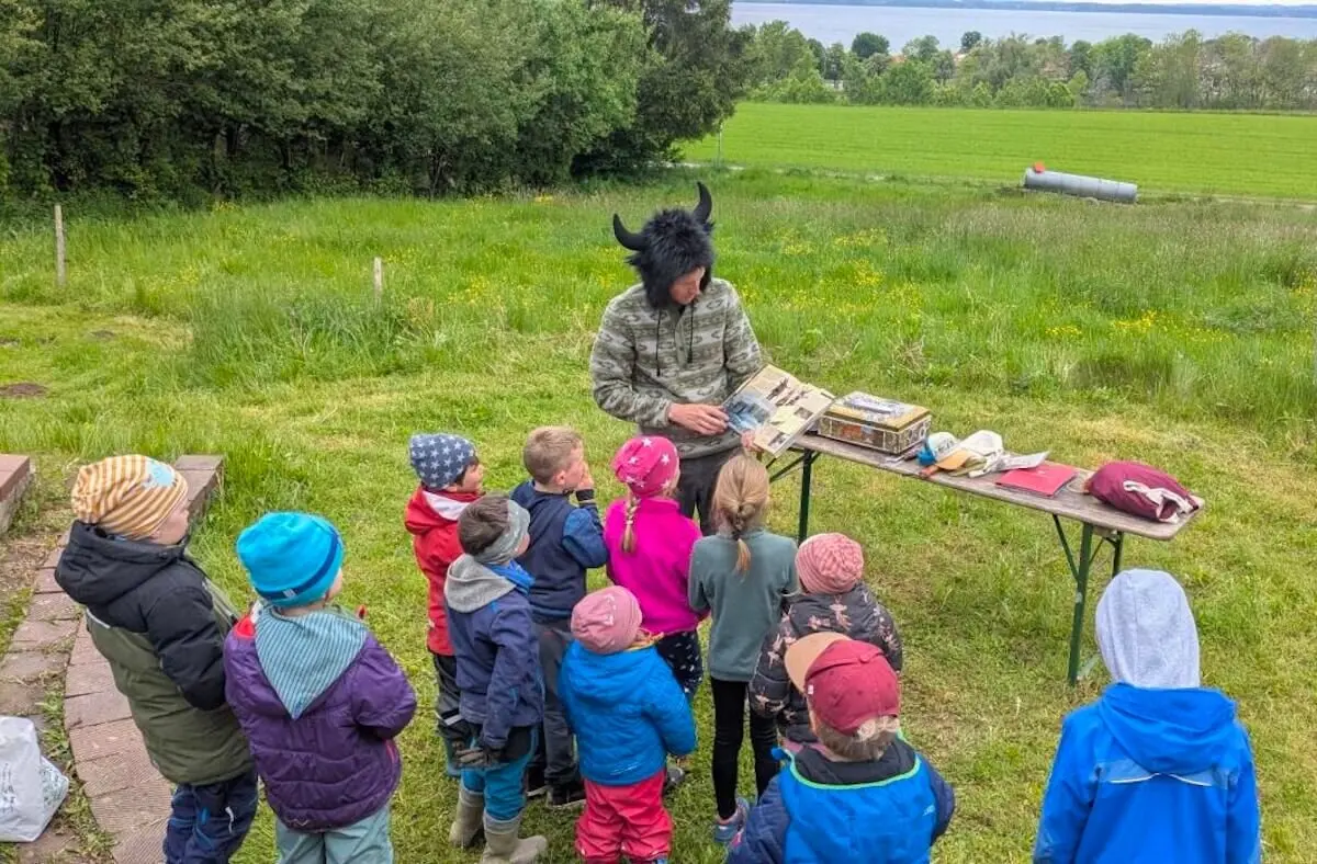 Eine Person mit einem Hut mit Hörnern zeigt einer Gruppe von Kindern draußen ein Buch. Sie stehen um einen Tisch mit Materialien. | © DAV Markt Schwaben | Foto: Familiengruppe