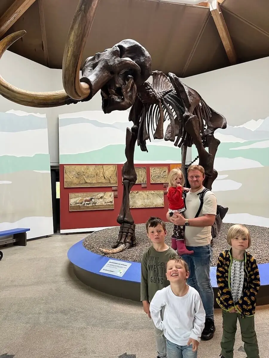 Eine Gruppe von Menschen steht vor einem großen Mammutskelett in einem Museum. Das imposante Skelett mit seinen langen Stoßzähnen ist auf einer erhöhten Plattform ausgestellt, umgeben von Fossilien und einem Wandgemälde mit Landschaftsmotiv. | © DAV Markt Schwaben | Familiengruppe