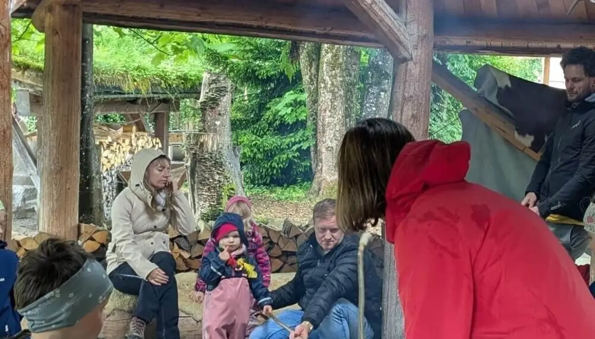 Eine Gruppe von Menschen, darunter Kinder und Erwachsene, versammelt sich unter einer hölzernen Überdachung im Wald. Einige halten Stöcke über ein Feuer, während im Hintergrund ein Stapel Brennholz zu sehen ist. Die Szene vermittelt eine gemütliche und gemeinschaftliche Atmosphäre bei einem Outdoor-Erlebnis. | © DAV Markt Schwaben | Familiengruppe