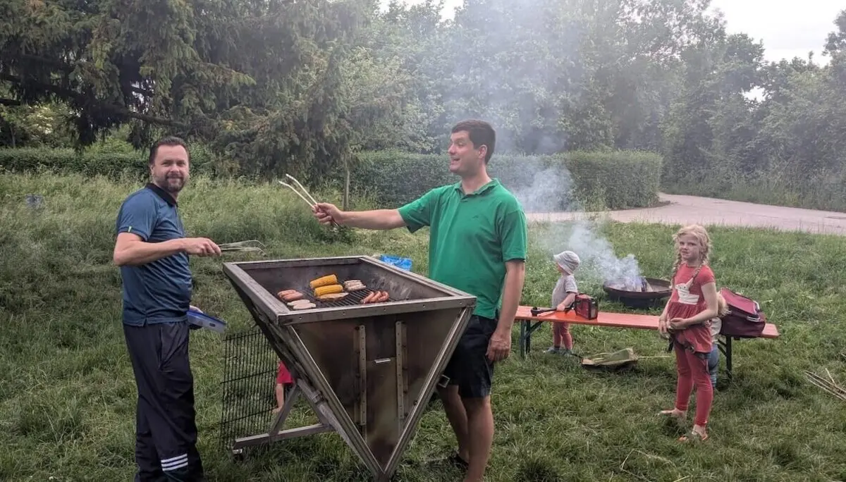Zwei Erwachsene grillen auf einem großen Grill Fleisch und Gemüse, Kinder stehen in der Nähe – entspanntes Beisammensein im Grünen. | © DAV Markt Schwaben | Foto Silke Titze