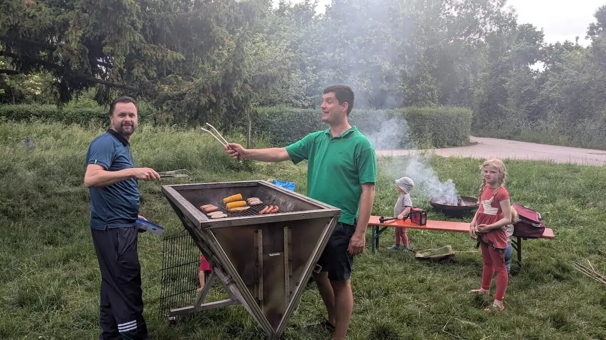 Zwei Erwachsene grillen auf einem großen Grill Fleisch und Gemüse, Kinder stehen in der Nähe – entspanntes Beisammensein im Grünen. | © DAV Markt Schwaben | Foto Silke Titze